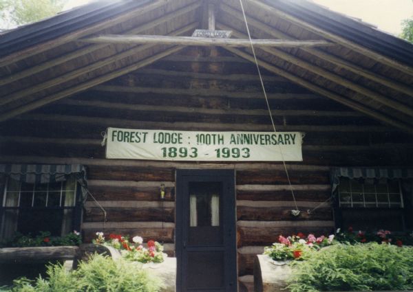 The front entrance to the main lodge with a banner above the door that reads: "FOREST LODGE : 100TH ANNIVERSARY 1893-1993." The lodge is constructed of chinked-horizontal logs with a log overhang. Flowers are in log planters on either side of the door and in log window boxes under the windows.