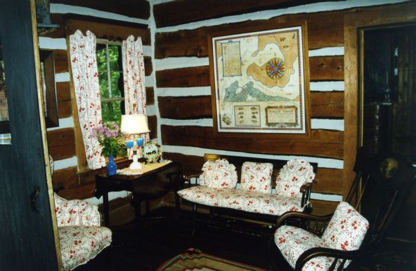White-chinked log walls with a framed map of Forest Lodge property. Dark wood furniture including a rocking chair, bench, side table, and an upholstered chair, all with red and white floral pads, plus matching curtains. A doorway leads to another room.