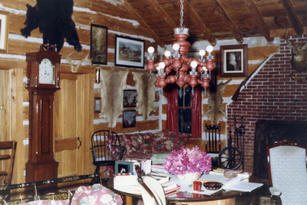A white chinked horizontal-log walls and plank-wood ceiling room that has a large brick fireplace with a deer mount near the top, a long-rifle in the center, and a spinning wheel and fireplace tools on the hearth. A chandelier hangs over an untidy table with a bouquet of flowers in the center. Animal skins hang on the walls along with numerous pictures and portraits. A grandfather clock sits against a wall between two doors, and chairs and benches are arranged around the room.