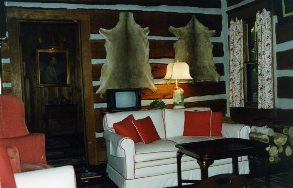 Horizontal white-chinked log walls with two mounted animal skins. A white sofa with red piping and red pillows is positioned in front of a table with a small television and a maiden-shaped porcelain lamp. A wood coffee table is in front of the couch and a stack of firewood is to the side. Floral curtains cover the window. A portrait of a young Mary Griggs is seen through the doorway.