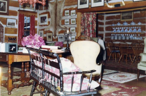 White-chinked horizontal log walls in living room looking into the dining room. An animal hide is hanging on wall and bull horns are mounted over the curtained doorway that looks into the dining room. Various rugs are scattered on the floor, and many pictures are hanging on the walls. A wood two seat bench with a back, arms and cushions faces the dining room. Chairs and two tables with books, flowers, and lamps are in the room. The dining room has a sideboard with a variety of dishes arranged on the shelves and a table with chairs.
