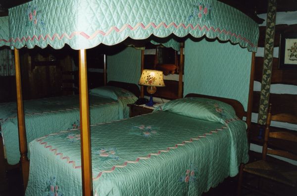 Two canopy twin beds with green turquoise quilted and embroidered canopy's and bedspreads. A wood chair with a woven seat is next to the bed as well as a tapestry embroidered bell strap hanging on the wall.  A small bed lamp is on a table between the two beds.