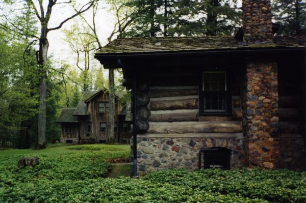 Rear view of horizontal log Main Lodge with cedar shake roof, stone chimney and foundation, surrounded by pachysandra and some lawn; the Guest House is in the background. Many trees, mostly pines, tower over the landscape.