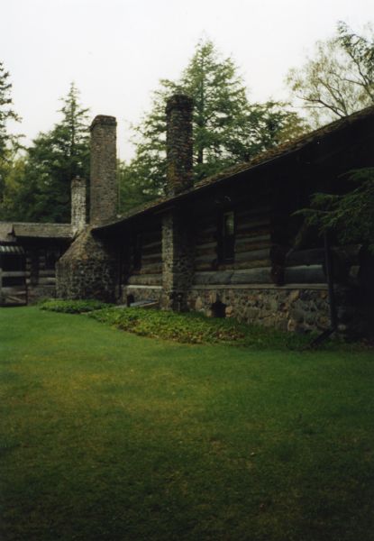 Horizontal single-story log home with cedar roof and stone foundation and three chimneys. A mowed lawn and foundation plantings surround the cabin.