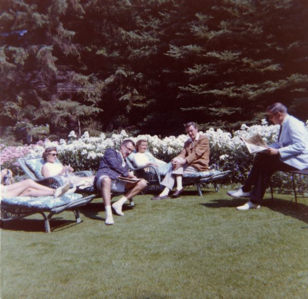 Mary and Jackson Burke are sharing a wicker chaise lounge on the lawn along with other couples sitting on wicker chaise lounges. The lawn is bordered by a white flower garden with very mature evergreen trees in the background. The women are wearing summer dresses or shorts, the men are dressed in sport jackets and slacks except one man is wearing tennis shorts and shoes.