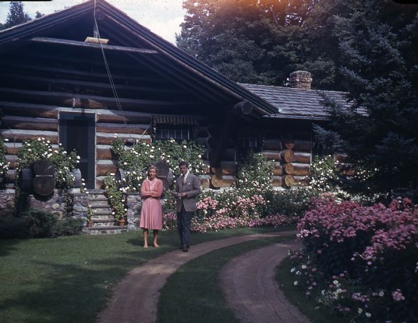 Mary Griggs, wearing a salmon colored dress and cardigan is standing in front of the Main Lodge with a male friend who is wearing a gray suit and tie while smoking a cigarette. There is a profusion of pink and white flowers growing all along the front of the lodge. A two rut dirt lane curves in front of the horizontal log lodge with a stone porch and foundation.