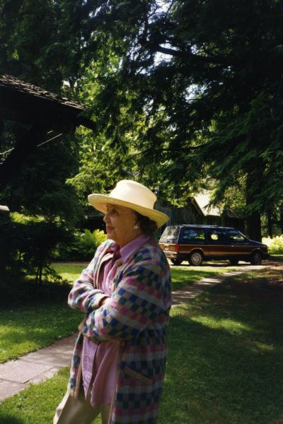 Mary Griggs Burke standing with her arms crossed in the yard by the main lodge under evergreen trees during the 100th anniversary celebration for Forest Lodge. Mary is wearing a long cardigan sweater, tan pants, a pink shirt and a straw hat. A minivan is parked in the background.