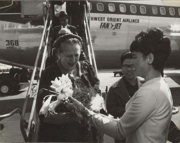 American journalist Esther Van Wagoner Tufty arriving in Seoul, Korea, at Kimpo (Gimbo) Airport, on a Northwest Orient Airlines airplane. Captions on album page reads: "arrive at Kimpo airport" and "Staff of Yowon present flowers". Because of her signature braided hairstyle, Mrs. Tufty was often referred to as "the Duchess."