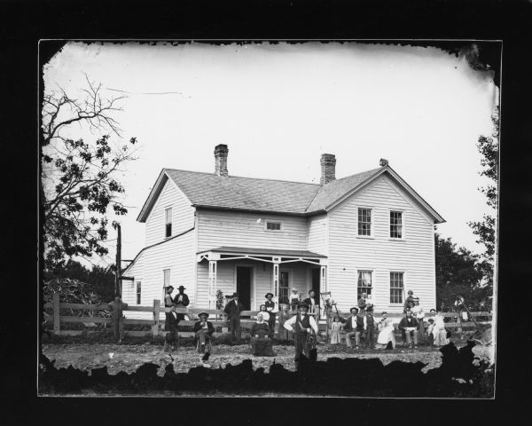Twenty-eight people and one dog are posed in the yard of a wood frame house. A lap dog is sitting on a chair balanced on top of a fence post. A woman at the far right is spinning. An elderly man is smoking a pipe. Three women are holding infants.