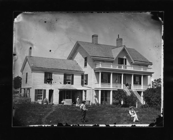 Allen E. Adsit is in the left foreground in this photograph and his father, Stephen Adsit, is seated at right. Seen near the house are a wooden rain barrel, a canopied well and grindstone. A woman is standing near the porch on the left, and people are on the second-story porch on the right.