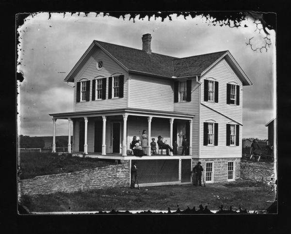 A family is seated on the wrap-around porch, with two men posed holding shotguns standing below them at foundation level. There are two windows in the stone foundation at the right. A stone wall is along the front yard on the left. In the background on the right is a man sawing wood with a buck saw, while a boy works next to him with an axe. The two-story frame house has a deep stone foundation, an octagonal window at the front of the house on the left, and full window shutters. One man seated on the porch is holding a document so that it can be plainly seen by the camera.