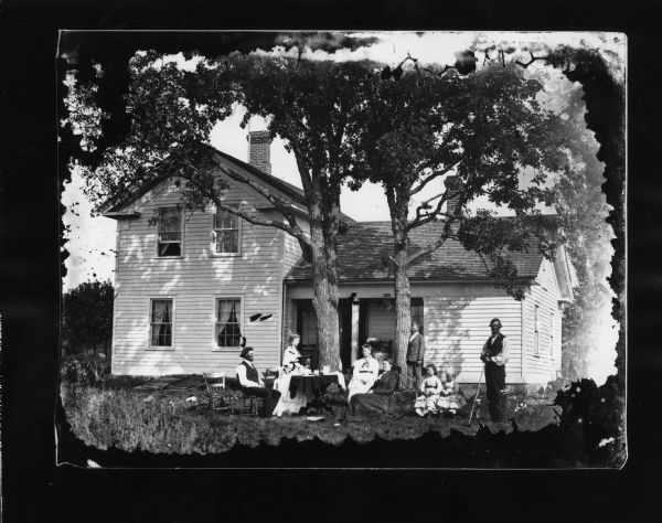 Family in yard around table, and a man standing with tool on the right. Behind the group is a Greek Revival house that has a sewing machine on the porch. There is a Hekla sign above the porch, and a cap is in tree in front of house.