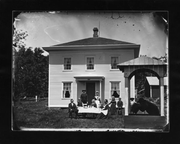 Here Dahl used "The Old Oaken Bucket" motif — a man drinking water from a pail at a well in foreground with family sitting and standing at a table with coffee behind. There is a frame house with Hekla Fire Insurance sign above door behind the farmily. There is a split-rail fence in the background.