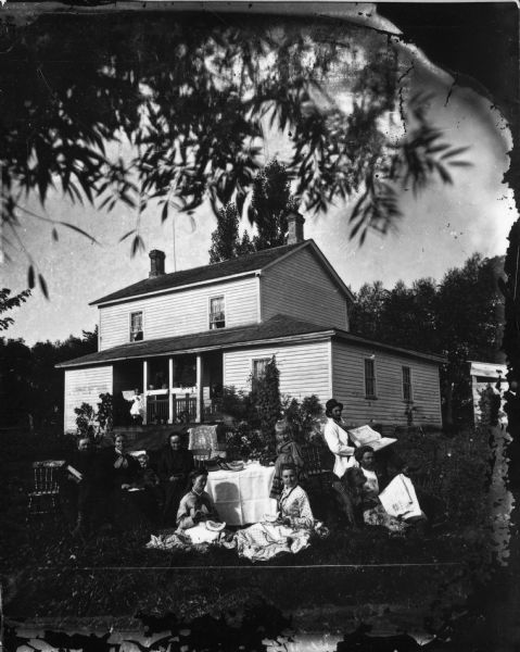 The Skavlem family is around the table at the home of Lars H. Skavlem.  A cloth, books, a vase of flowers and fruit are on the table.  The two men on the right have pipes. One has the "Combination Atlas Map of Rock County" (1873); the other has the May 18, 1875 edition of "Skandinaven," a newspaper published in Chicago. The man in the right rear is probably Halvor L. Skavlem. Two women in the front have watermelons. The woman on the front left is probably Halvor's wife, Gunil Olmstad Skavlem. The man and woman on the left are Lars H. Skavlem and Groe Halvorsdatter Aae. Two chairs at the table are empty. A girl is sitting at an organ on the porch, which has many plants on it. A lightning rod is on the roof.
