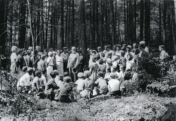 Soren Uhrenholdt is holding his hat as he stands amongst a group of 4-H children and young adults in a Sawyer County pine forest.