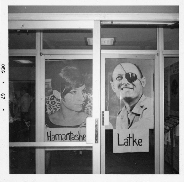 A view of the front doors of the Hillel Foundation, with posters of Barbara Streisand and Moshe Dayan. There are signs under each poster: "Hamantashe" for Streisand, and "Latke" for Dayan. 
