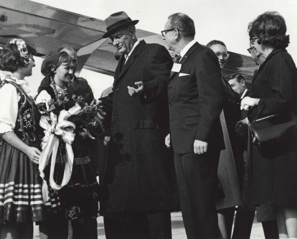 President Lyndon Johnson accompanied by Governor John Reynolds and Senator Gaylord Nelson is greeted at Mitchell International Airport by girls dressed in Polish garb while on his 1964 Presidential campaign. One young lady presents flowers and another, Barbara Banasajowski, presents a sheaf of wheat, a symbol of a bountiful harvest.