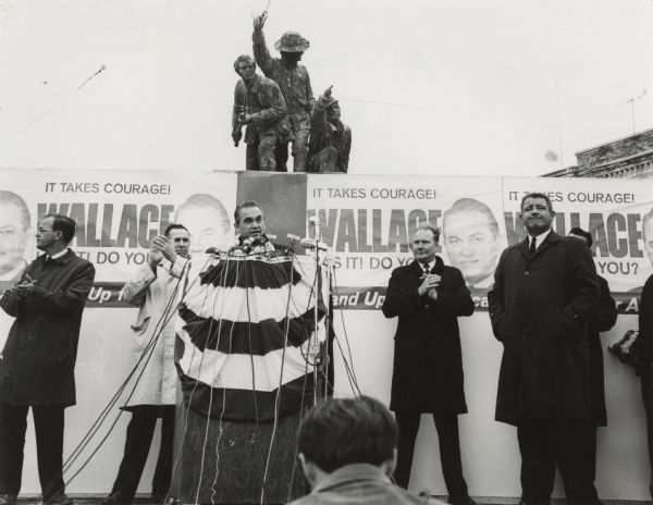 George Wallace stands at a podium with bunting and several microphones during a rally for his Presidential primary campaign in Oshkosh. Five other unidentified men are standing nearby, some applauding in support. Posters with Wallace's portrait are behind the stage which read: "It takes courage! Wallace has it! Do you? Stand Up for America!". The Oshkosh Civil War monument is visible above the wall of posters behind Wallace.