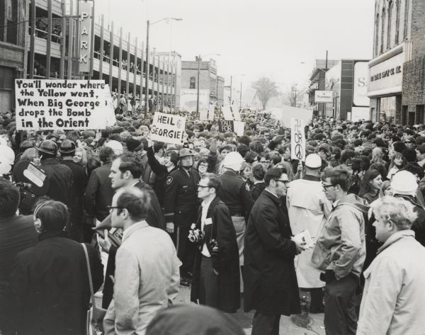 A large group of people, many carrying signs opposing George Wallace, gather in downtown Oshkosh near a Wallace rally. Some of the signs read: "You'll wonder where the Yellow went, When Big George drops the Bomb in the Orient", "Heil Georgie!". Another sign has a symbol of the swastika, the word "Or" and the peace sign. A man in the foreground has two cameras around his neck, and a television camera on the left has a label on it that reads: "BBC-TV".