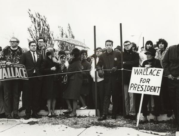 Supporters of Alabama Governor George Wallace stand behind a rope at a Wallace rally during the 1964 Presidential primary campaign. One woman is holding a sign that reads: "Wallace For President". Another man has a "Wallace" sticker on his forehead.