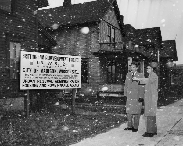 Two men stand on a sidewalk and look in the direction of a sign, which reads: "Brittingham Redevelopment Project UR Wis. 2-1 A Project of City of Madison, Wisconsin  This project is undertaken with federal financial aid under Title I of the Housing Act of 1949 as amended through the.. Urban Renewal Administration Housing and Home Finance Agency." A caption on the rear of the image identifies the man on the left as Mayor Ivan Nestigen.