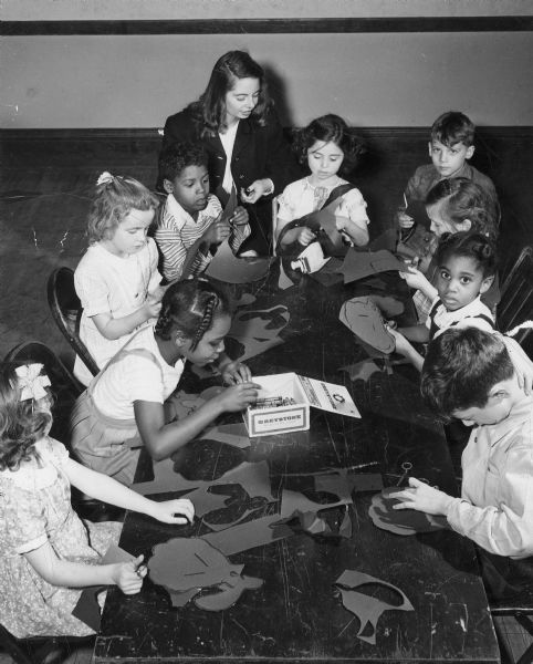 Slightly elevated view of nine children sitting around a table working on craft projects. A woman is sitting with them at the top of the table.