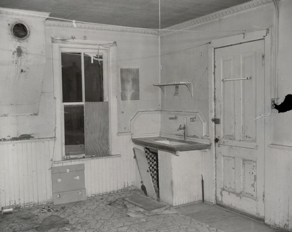 Interior view of a room. The window on the far wall is partially boarded up. The cabinet door under the sink is ajar, the floor has broken or missing pieces, and general damage and wear and tear is apparent throughout the room.