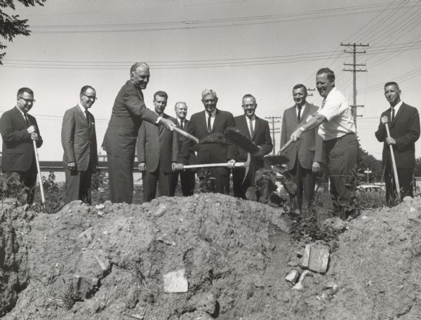 Ten men are standing near a pit, which is in the foreground. All but one of the men are wearing suits, and several are holding shovels or pickaxes. Two men are tossing the dirt from their shovels.