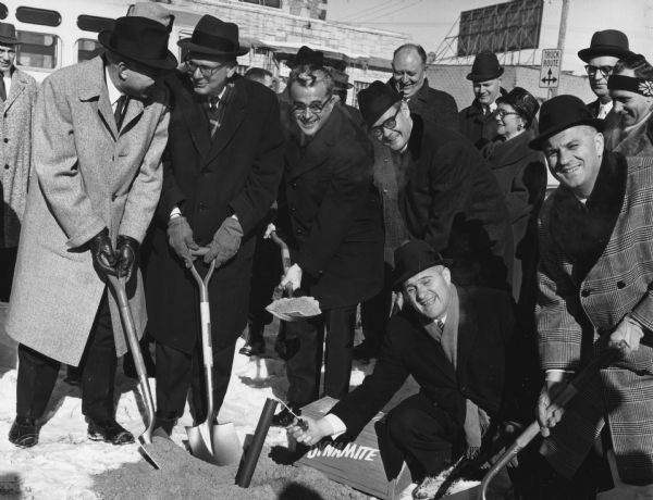 Six men are posing around a patch of dirt, and snow is on the ground. Five of the men are holding shovels, and one man is lighting what appears to be a prop stick of dynamite. A crate behind it is labeled: "DYNAMITE." Other men and women are looking on from the background.