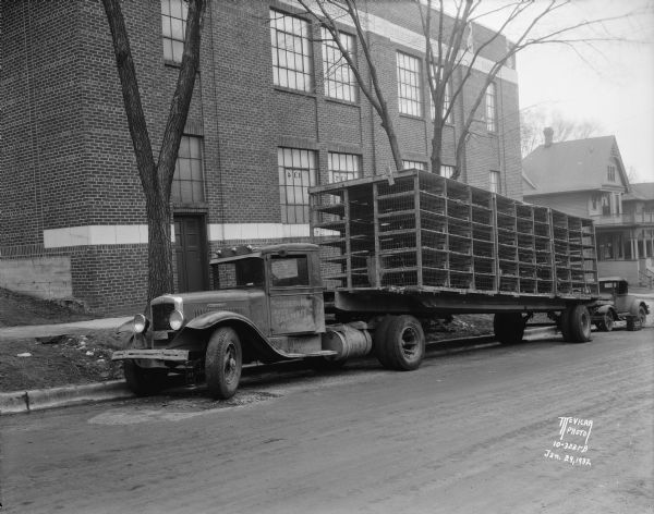 View across street towards an International truck with poultry rack trailer parked along a curb. The sign painted on the driver's side door reads: "Southern Minn Auto Transit Mankato". An automobile is parked behind the truck, and a box, perhaps for cameras, is sitting on the street near the driver's side door and the front of the box reads: "M<u>c</u> Vicar Photo Service".