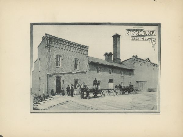 View of the George Ruder Brewing Company. A group of men and a dog are posing near an entrance on the side of the brick building. A man is on a horse-drawn wagon, and another horse-drawn wagon is behind him on the right.