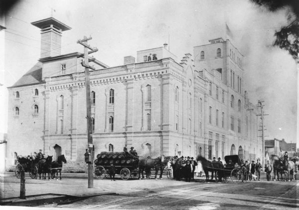 View down street towards the George Walter Brewing Company. A large group of people are posing on the street with horse-drawn carriages and horse-drawn wagons loaded with barrels of beer at the corner of the building. A man in the center of the group is holding up a broom. Two people are standing on the roof looking down at the group.