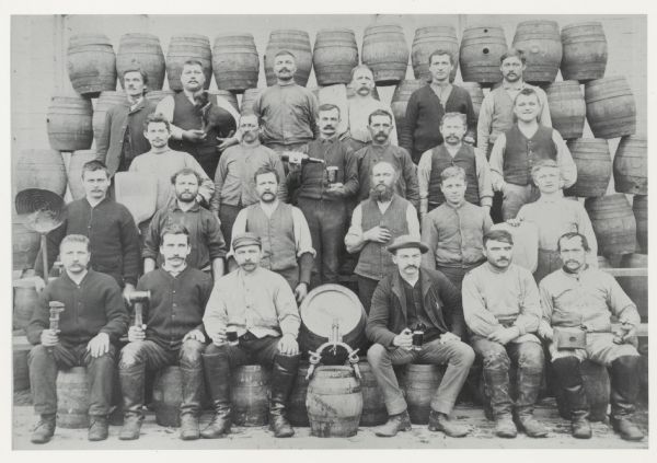 Brewery workers, posed in front of stacked beer barrels. Pictured (2nd row, far right) is Louis Schreiber (1861-1923) who immigrated to Wisconsin from Bavaria at age 18. The man in the top row, second from left, is holding a dog in his arms. The tapped barrel is marked John Gund Bottling and the barrel at front center is stamped "J.GUND BO."