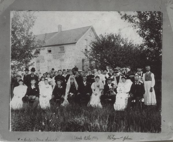 A large group is posing sitting in the yard of a stone building on the day of the wedding of Bertha Wendtland and William Klug. Some of the people in the group are standing and holding plates of food, and others are holding glasses while a man is pouring something from a bottle. Below the photograph are handwritten: "&#8730; = Pastor and Mrs Seyel(?)"; Uncle Willie King(?)" and "Netgows place(?)". There are &#8730; marks above a man on the left, and a woman in the center near the bride. 