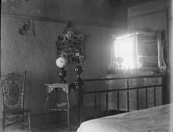 Interior of a bedroom. In the corner is a dresser with a mirror, a bed is in the foreground, and on the left are a side table with a lamp, and a chair. There is a collage of photographs hanging on the wall.