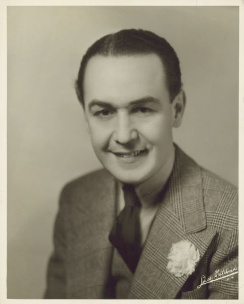 Head shot of actor Joseph Downing. He looks straight at the camera and smiles.  Downing wears a suit coat, vest, tie and a carnation in his lapel.