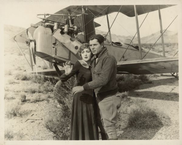 Warner Baxter and Billie Dove stand in front of an airplane in a scene from the 1925 film The Air Mail.  They are in a half-embrace and both have an arm up to defend themselves.  