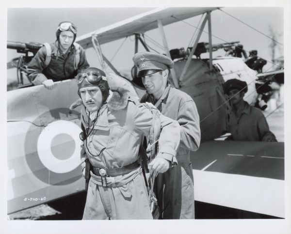 A man helps Jack Holt put on a parachute in a scene from the 1935 film Storm Over the Andes.  Two men in the background, one in a plane and one standing by the wing, look on.