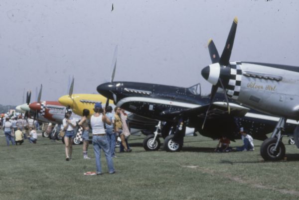 Visitors to the Experimental Aircraft Association (EAA) AirVenture fly-in are looking at several airplanes lined up on the airfield.