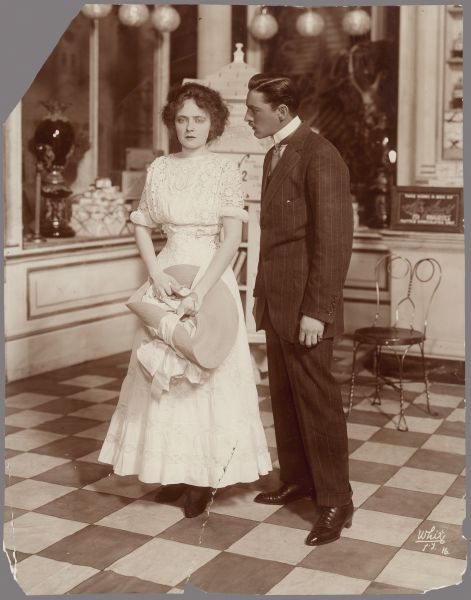 John Barrymore and Eda Bruna stand next to each other in a scene from the 1909-1910 play The Fortune Hunter.  They appear to be in a restaurant or a drug store.  Barrymore looks intently at Bruna who looks straight ahead.