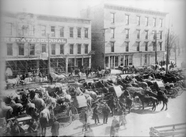 Elevated view of the farmers' market at the base of the water tower (not in view) in front of the Washington Building. There is a Wisconsin State Journal sign at the 100 block of E. Washington Avenue. The farmers are standing with their horses and wagons, with many parked in the center of the street. A large sign on the building on the left reads, in part: "IN STATE JOURNAL". Signs on the storefronts for the buildings on the left read: "Piper Bros Groceries and Seeds", " J. P. Halbach" and "Cook Bros". A sign on the building on the right reads: "Conklin & Sons Coal, Wood & Ice". 