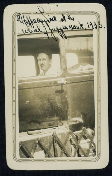 Ed Hoffmann Price is pictured at the wheel of his automobile the "Juggernaut". A metal gate along the running board is holding in a suitcase and a can of "Cross Country Motor Oil" against the driver's side door.