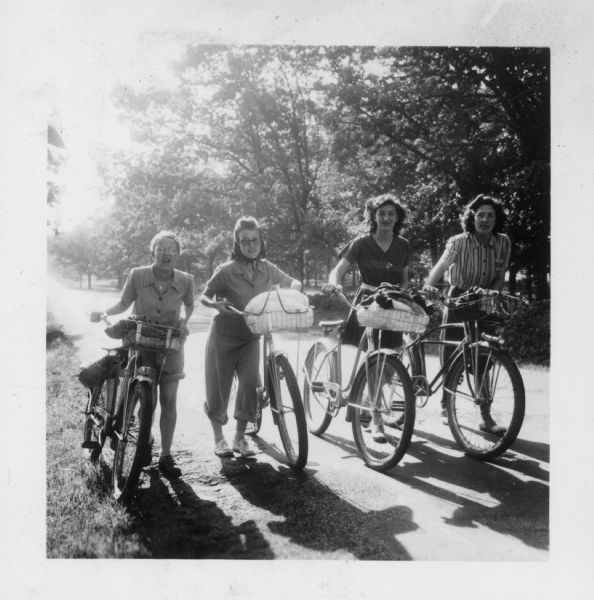 Gail Taliaferro, Florence Vivian, Catherine Cefalu, and Charlotte Barber standing with their bicycles, which are loaded with supplies, on a tree-lined road for the 4th Neighborhood House American Youth Hostel group bicycle trip to the Cross Plains Hostel.