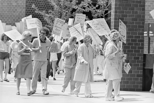 Men and women picketing outside a hospital, likely UW Hospital. Some of signs they are carrying read: "Poor Staffing is Dangerous to Your Health", "Citizens UP of UP Wisconsin Deserve Health Care", and "Nurses are an Endangered Species! Save the Nurses!!"