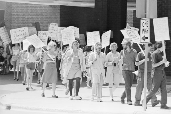 Men and women picketing outside a hospital. Some of signs they are carrying read: "Hospital Workers Together!", "Nurses An Endangered Species UP" and "Wisconsin Deserves Quality Health Care But: You Can't Run a Hospital Without S[taff] UP/1199".