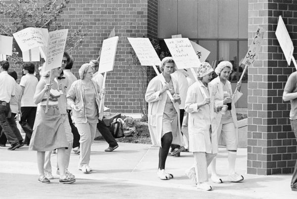 Men and women picketing outside a hospital. Some of signs they are carrying read: "Poor Staffing is Dangerous to Your Health", "Wanted!! Cheap labor Apply State of Wisconsin UW Hospitals" and "UWH Staff is Dedicated Not Donated".