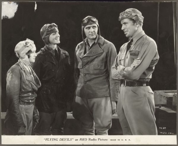 Still from the 1933 RKO film Flying Devils.  Arline Judge, Eric Linden, and Ralph Bellamy stand next to each other and look at Bruce Cabot who looks directly at Judge.  They are all wearing, uniforms, flight caps and goggles.