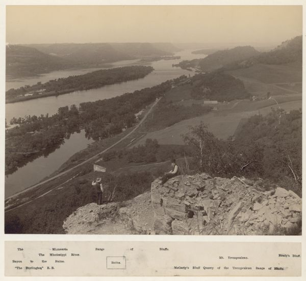 Elevated view of two men on a rock formation in the foreground, one man sitting, and one man standing holding a flag. Below them is the Mississippi River and bluffs on the opposite shoreline. Site of Perrot's wintering post, 1885-1886. The label below the photograph identifies: The Minnesota Range of Bluffs, The Mississippi River, Bayou to the Ruins, Ruins, The Burlington R.R., Mt. Trempealeau, Brady's Bluff, and McCarty's Bluff Quarry of the Trempealeau Range of Bluffs.