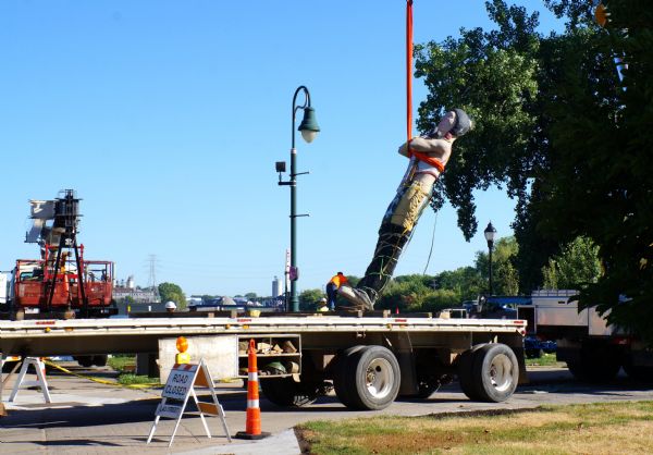 A man is standing on top of a trailer loading a large statue of a Native American man attached to a crane.
