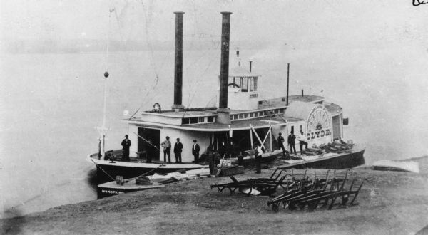 The sidewheel steam rafter "Clyde," which was the first iron hull steamboat. Sign on side of boat reads: "Dubuque, Reads Landing & Eau Claire." Agricultural implements, plows, are lined up along the shoreline in the foreground.